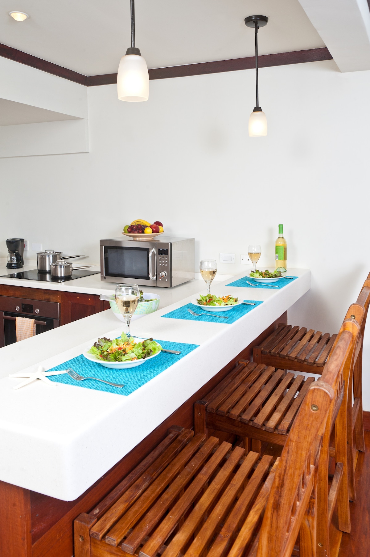 A modern kitchen space features a white counter bar with three wooden stools. Two plates of salad and glasses are positioned on bright blue placemats. Above, pendant lights provide soft lighting, while a microwave and coffee maker are visible in the background.