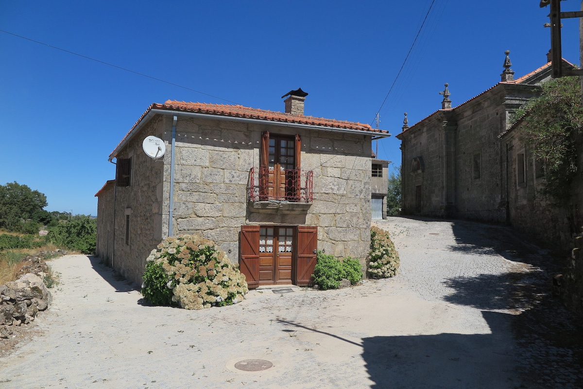 A rustic stone house with a red-tiled roof is situated on a cobblestone street. A small balcony with decorative ironwork overlooks the entrance, which features wooden doors. Surrounding vegetation adds a touch of greenery to the rural setting, with clear blue skies overhead.