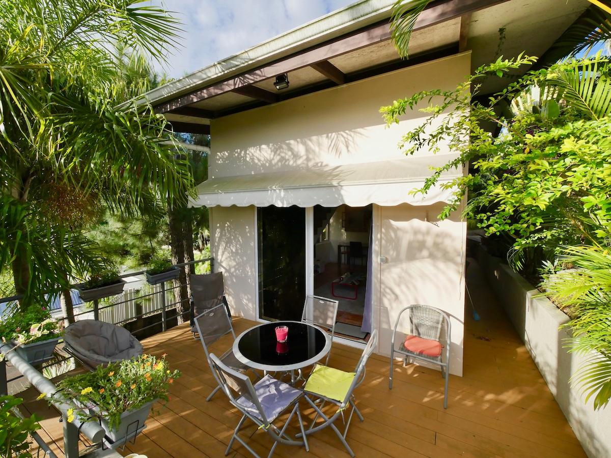 An outdoor terrace is surrounded by tropical foliage, featuring a circular table with six chairs. A small plant arrangement adds a touch of greenery, while a retractable awning provides shade. Large glass doors lead into the living space, enhancing the connection to nature.