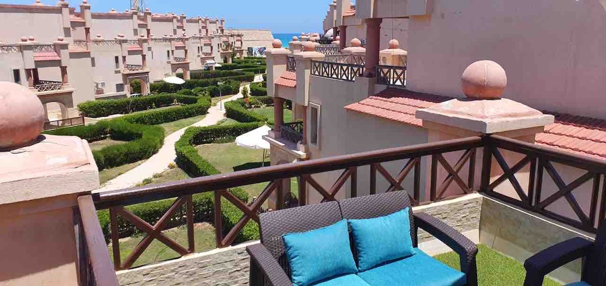 A balcony view showcases a wicker seating area with blue cushions, overlooking landscaped gardens and pathways. The distant sea is visible against a clear sky, providing a serene backdrop. Architectural details of nearby buildings add character to the scene.