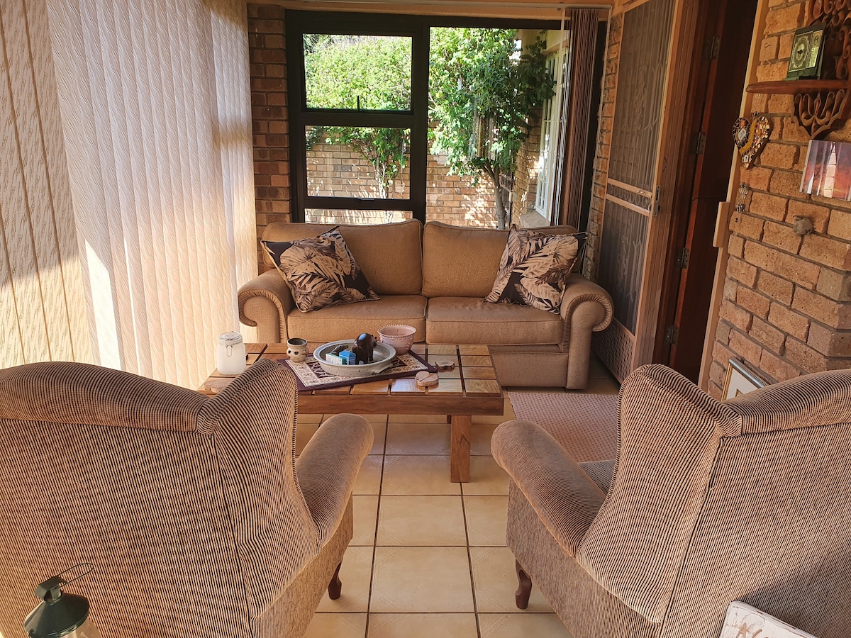 A sunlit sitting area features a comfortable sofa adorned with decorative pillows, complemented by two upholstered chairs. A wooden coffee table occupies the center, displaying a few casual items. Large windows bring in natural light and reveal greenery outside.