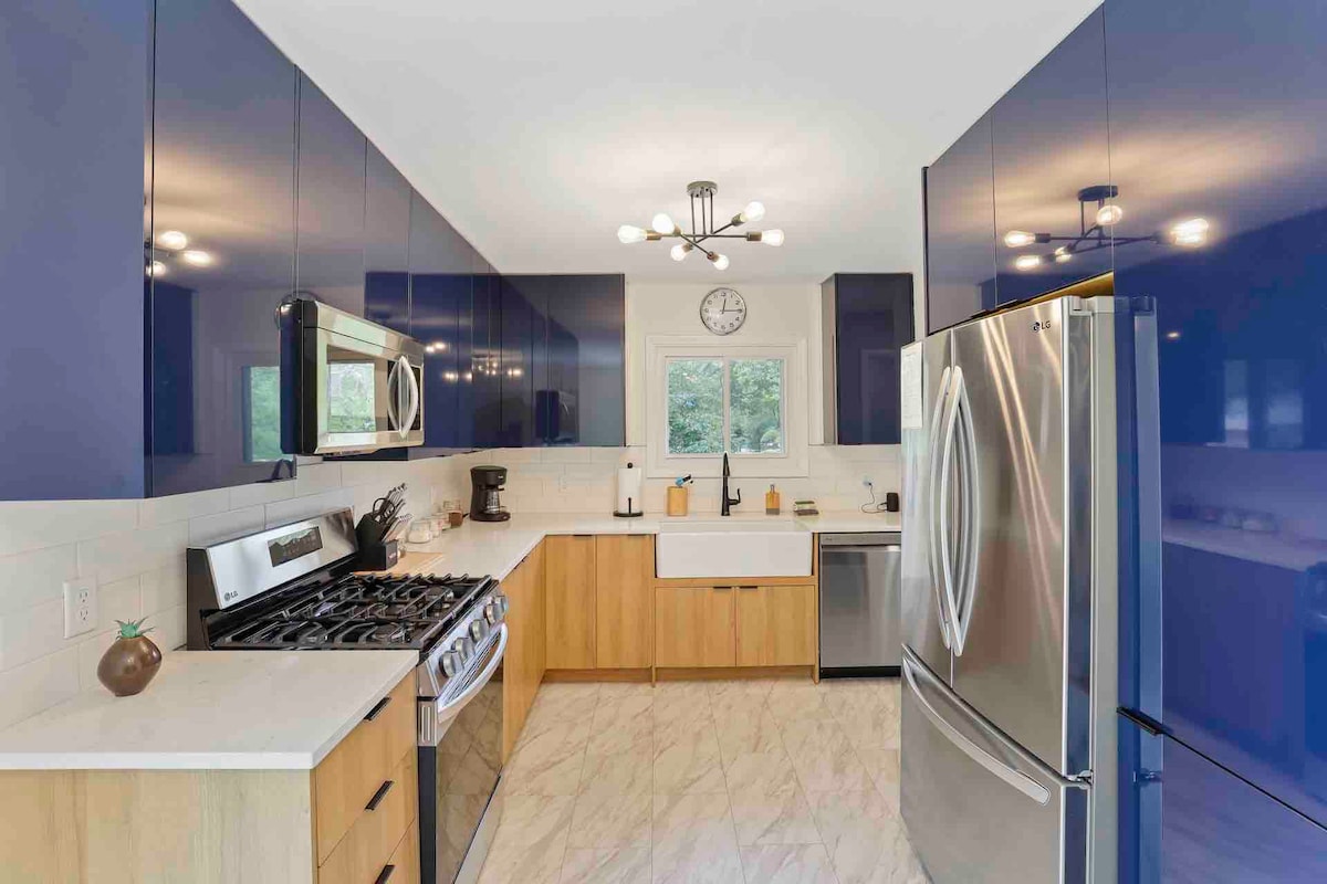The kitchen features contemporary blue cabinetry paired with light wood accents. A large stainless steel refrigerator stands beside a gas stove. A white farmhouse sink is centered beneath a window, while a modern light fixture illuminates the space.