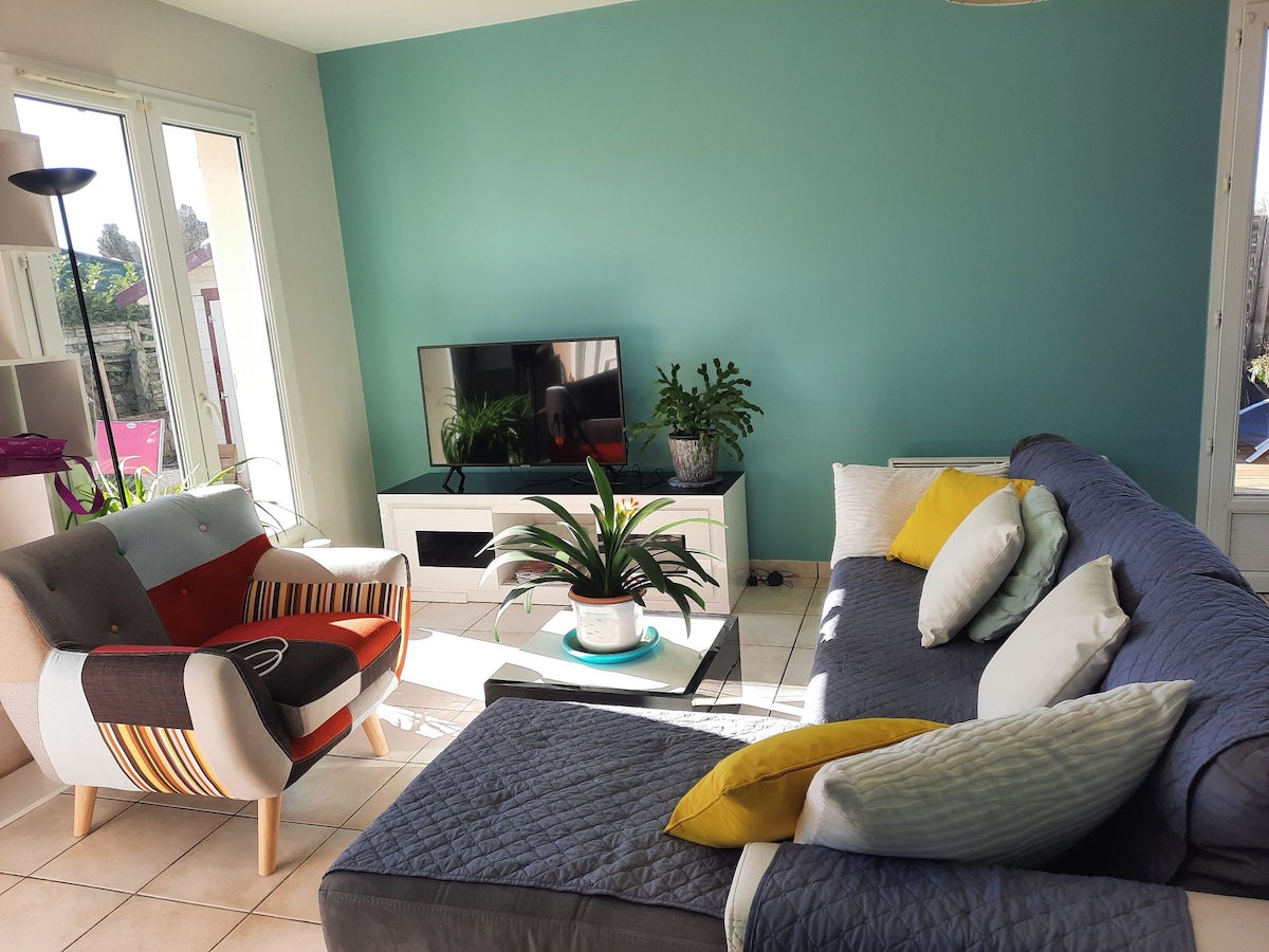 A bright living area features a comfortable sectional sofa adorned with various cushions. A small, colorful armchair complements the space, while a modern television is positioned on a low white media unit. Natural light flows in through large windows, enhancing the fresh wall color.