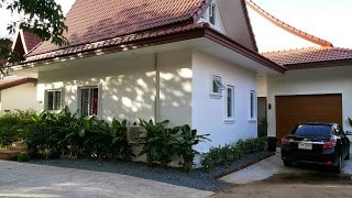 A white cottage with a red tiled roof is surrounded by lush green foliage. A gravel driveway leads to a brown garage door, with a parked black car positioned nearby. Windows are present on either side of the exterior, contributing to the welcoming design.