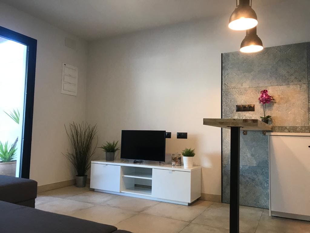 A modern living area is depicted, featuring a flat-screen television on a white console cabinet. Small potted plants are positioned nearby, and a minimalist bar area with pendant lighting is visible. Natural light brightens the space, highlighting the neutral tones of the walls and flooring.