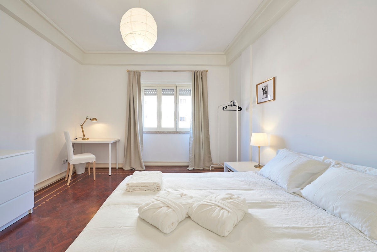 A well-lit bedroom features a spacious bed adorned with folded towels. A desk with a lamp and a chair is positioned beside a window, while a clothing rack stands nearby. Light curtains frame the window, and a soft pendant light hangs from the ceiling.