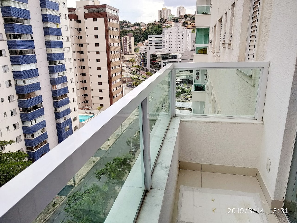 A balcony with glass railings offers a view of the cityscape. The floor is tiled, and the walls are painted in a neutral color. Surrounding buildings are visible, providing a sense of the neighborhood's urban environment. The area appears to be well-maintained and inviting.