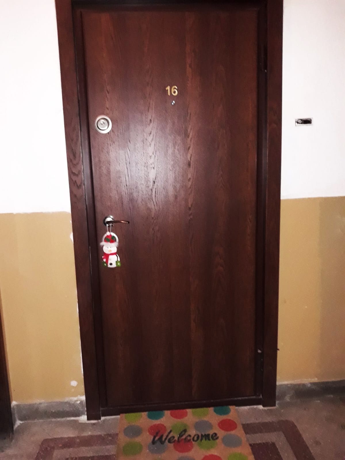 A wooden entrance door marked with the number 16 is situated at the end of a corridor. A welcome mat with colorful dots is placed in front, adding a friendly touch to the entryway. The wall displays a two-tone color scheme of light and dark beige.