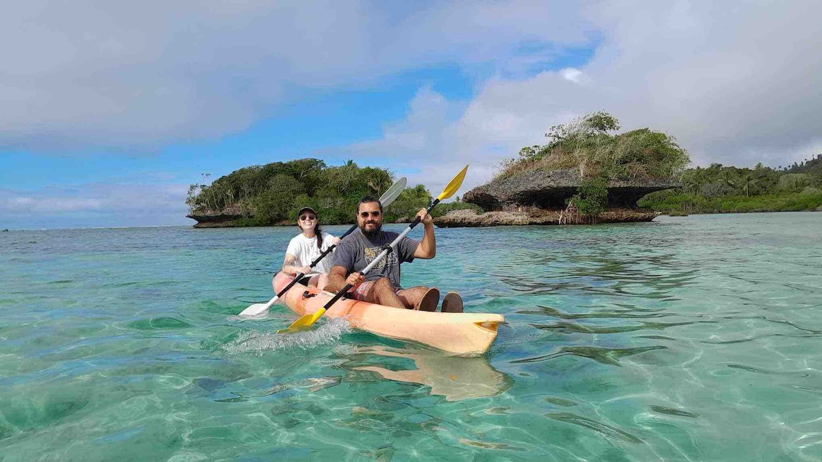 Two individuals are seen kayaking across clear, tranquil waters, surrounded by lush greenery and rocky formations. The kayakers appear engaged in their activity, with the vivid turquoise water reflecting the sky above. The serene landscape offers a sense of adventure and exploration.