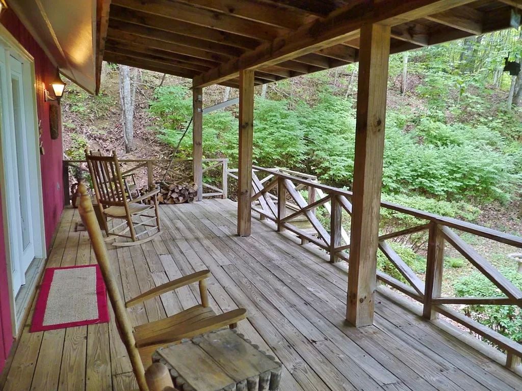 A large covered porch features wooden rocking chairs and a rustic table. The porch is surrounded by natural greenery, with a wooden railing offering an unobstructed view of the forested area below. A small welcome mat is positioned near the entrance.