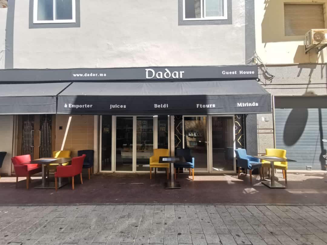 A modern guest house exterior is displayed, featuring a large awning with the name 'Dadar' prominently shown. Colorful outdoor seating options, including red and yellow chairs, accompany small tables. Large glass doors provide a view into the inviting interior space.