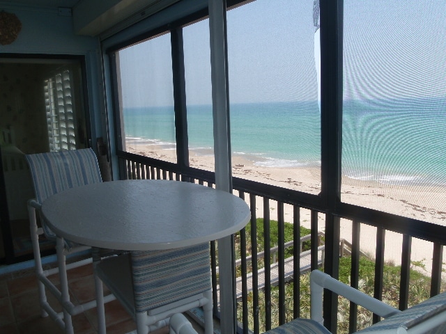A screened-in balcony offers a table with two chairs, overlooking a sandy beach and calm ocean waters. The view is unobstructed, allowing for a clear sight of the shoreline and distant waves. Light filters through the screens, creating a comfortable atmosphere.