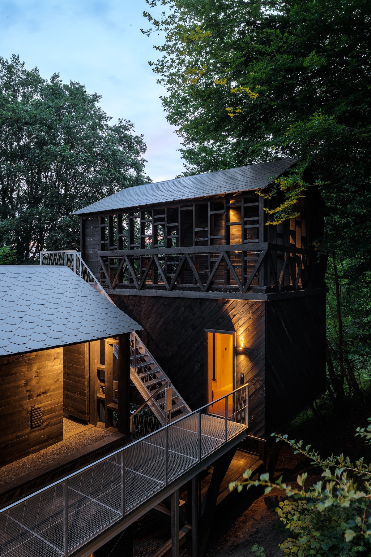 A multi-level wooden cabin is set among trees, featuring a covered balcony and outdoor staircase. Soft lighting accentuates the cabin's structure, highlighting its rustic appeal. The entrance door is clearly visible, leading into the inviting interior space.