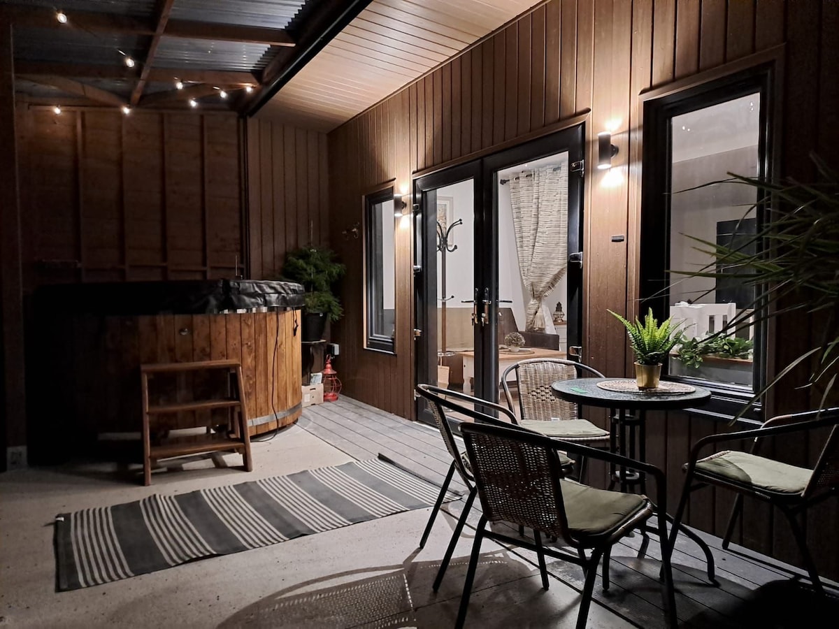 A private patio area features a wooden hot tub and a cozy seating arrangement with a small table and chairs. Soft outdoor lighting enhances the wooden walls, while potted plants provide a fresh touch. A patterned rug adds comfort underfoot.