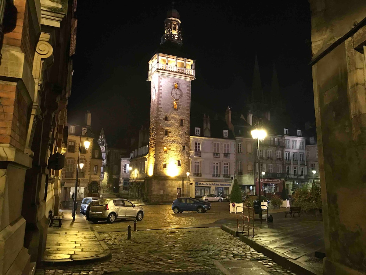 A historic clock tower is illuminated at night, casting a warm glow over the cobblestone square. Surrounding buildings display traditional architecture, with soft lighting enhancing the inviting atmosphere. Parked cars are visible, and several streetlights contribute to the serene setting.