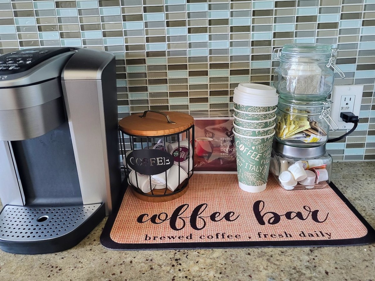 A coffee station is shown with a coffee maker positioned beside a woven basket containing single-serve coffee pods. A decorative mat labeled 'coffee bar' sits underneath, accompanied by stacked paper cups and various sugar and cream containers in glass jars.