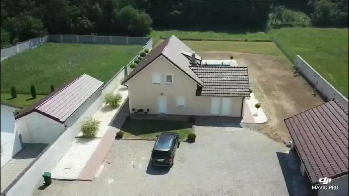 An aerial view of a spacious villa is shown, featuring a well-maintained lawn and surrounding greenery. The property includes a parking area with a vehicle, a patio accompanied by landscaping, and a private pool visible at the rear.