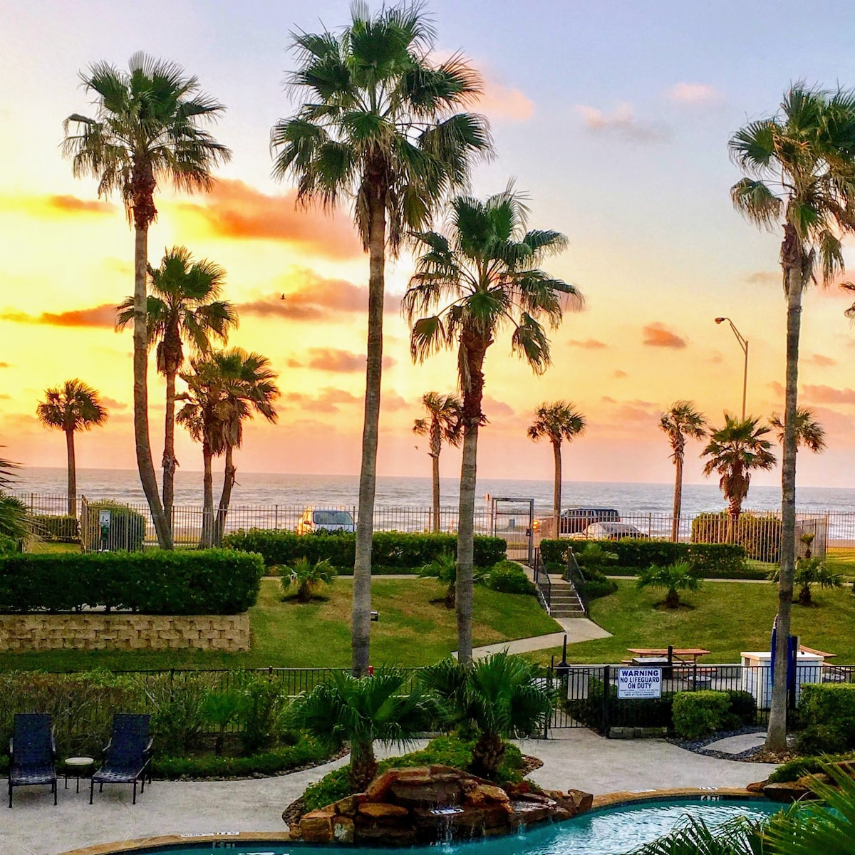 Palm trees frame a serene view of the Gulf of Mexico, where gentle waves meet sandy shores. A vibrant sunset casts warm hues across the sky, illuminating the nearby pool area and lush landscaping. The scene conveys a feeling of tranquility and natural beauty.
