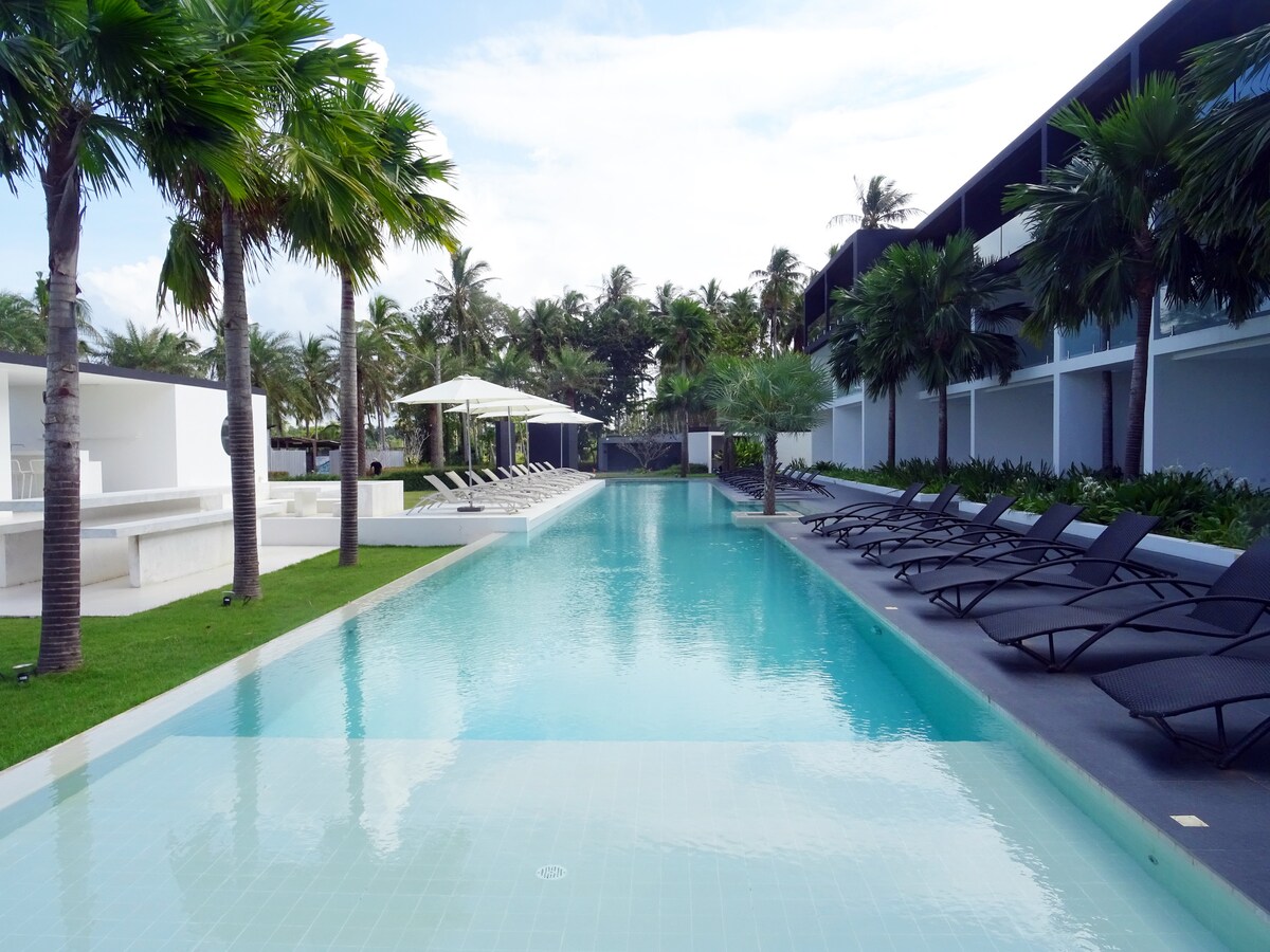 A long swimming pool is bordered by palm trees and lounge chairs. Sun loungers are positioned along one side, providing space for relaxation. The contemporary condominium building is visible in the background, framed by lush greenery and a clear sky.