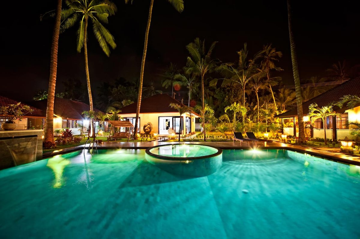 A tranquil nighttime scene displays a spacious swimming pool surrounded by lush palm trees. Soft lighting highlights the area, reflecting off the water. Several lounge chairs are positioned nearby, inviting relaxation, while charming tropical architecture frames the backdrop.
