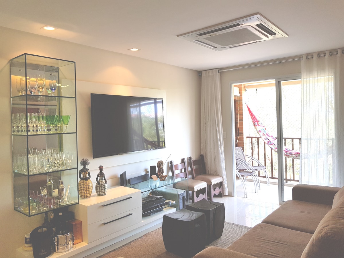 The living area is fitted with a large flat-screen TV mounted on the wall above a stylish white console. A glass display cabinet showcases decorative items and glassware. Natural light flows in from the balcony, which is furnished with a hammock and seating.