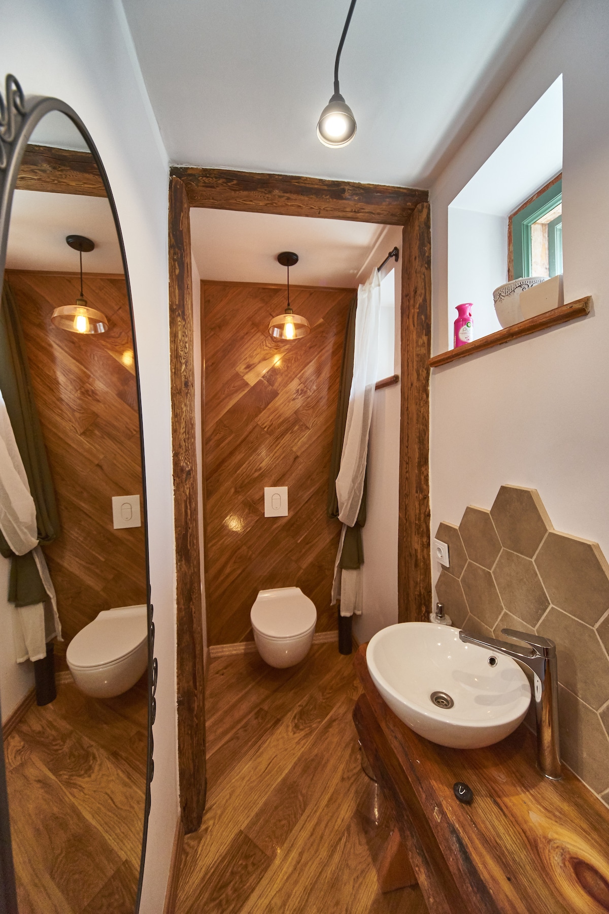 A modern bathroom is featured, showcasing warm wooden accents and geometric tiles. A circular mirror reflects the space, which includes a wall-mounted sink and a toilet. Natural light enters through a small window, enhancing the overall ambiance.