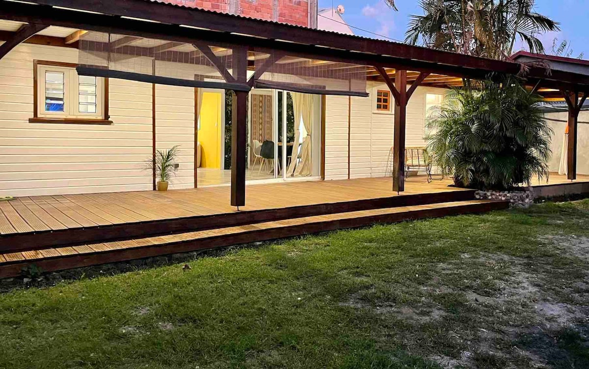 A spacious wooden deck extends from the house, featuring a covered area supported by dark wooden beams. Clear glass doors allow visibility into the interior. Lush greenery and a small potted plant are positioned along the deck, enhancing the connection to the outdoors.