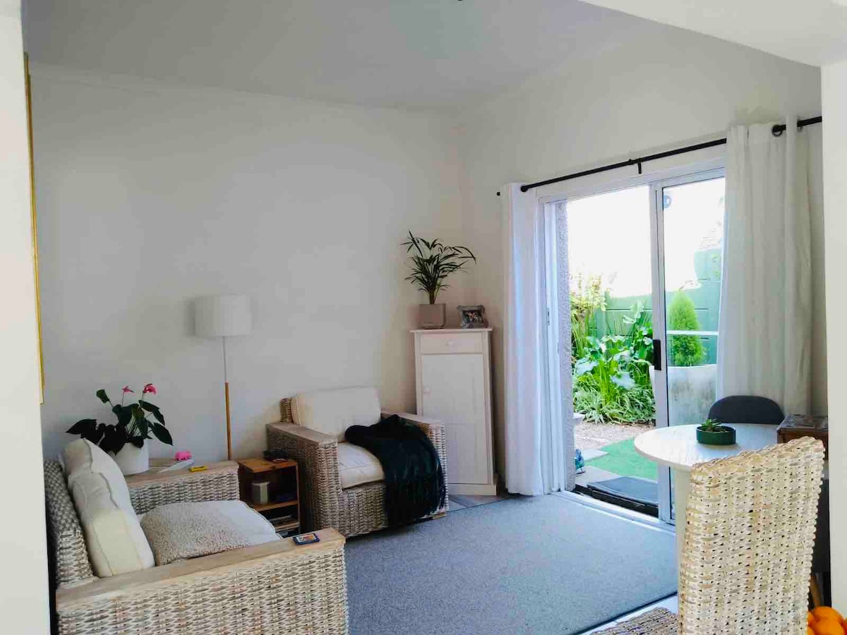 A bright, open-plan lounge area is furnished with natural wicker chairs and a sofa. Light curtains frame the sliding glass door, which opens to a garden view. A small dining table with a single chair sits beside the door, complemented by a potted plant.