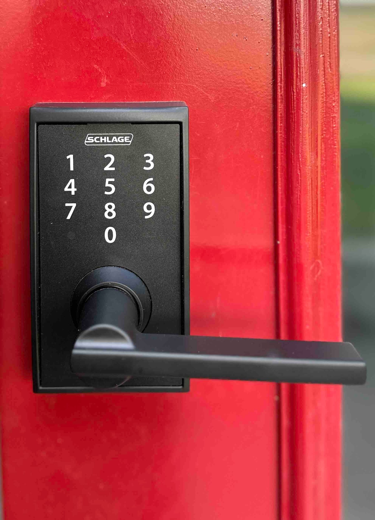 A close-up view of a black electronic keypad lock is displayed on a vibrant red door. The keypad features numbers 0 through 9 arranged in two vertical columns. A sleek, black lever handle is positioned to the right.
