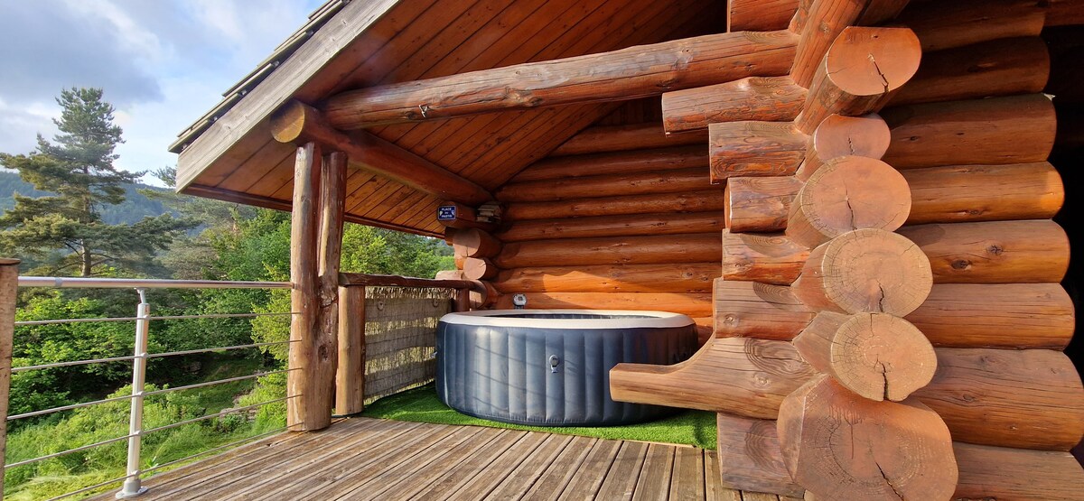 A log chalet features a private terrace showcasing a circular Jacuzzi, surrounded by natural greenery. The wooden structure is complemented by the rustic logs and a railing, providing a serene outdoor space for relaxation.