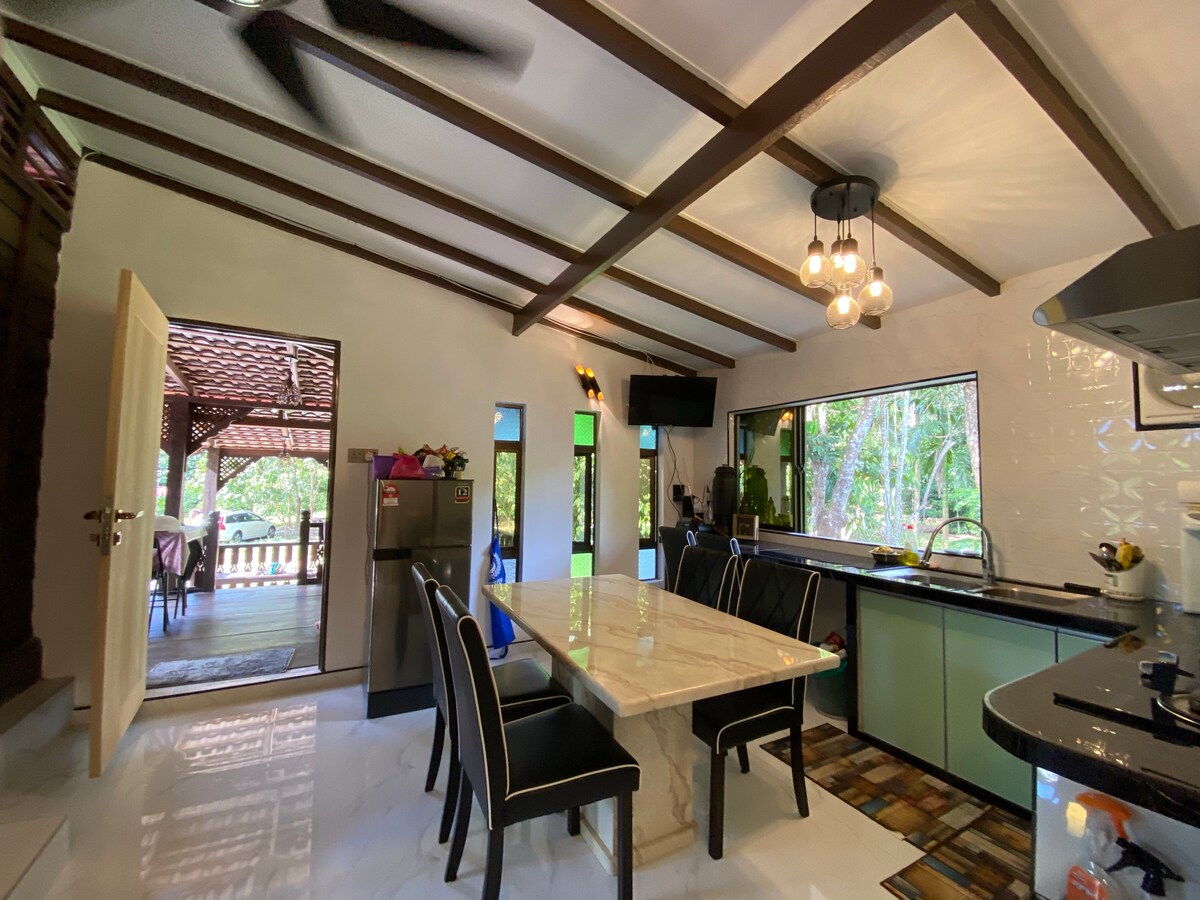 A bright kitchen area features a spacious marble dining table surrounded by black chairs. Large windows allow natural light to fill the space, and views of the surrounding greenery can be seen through the open doorway. The kitchen is equipped with modern appliances.