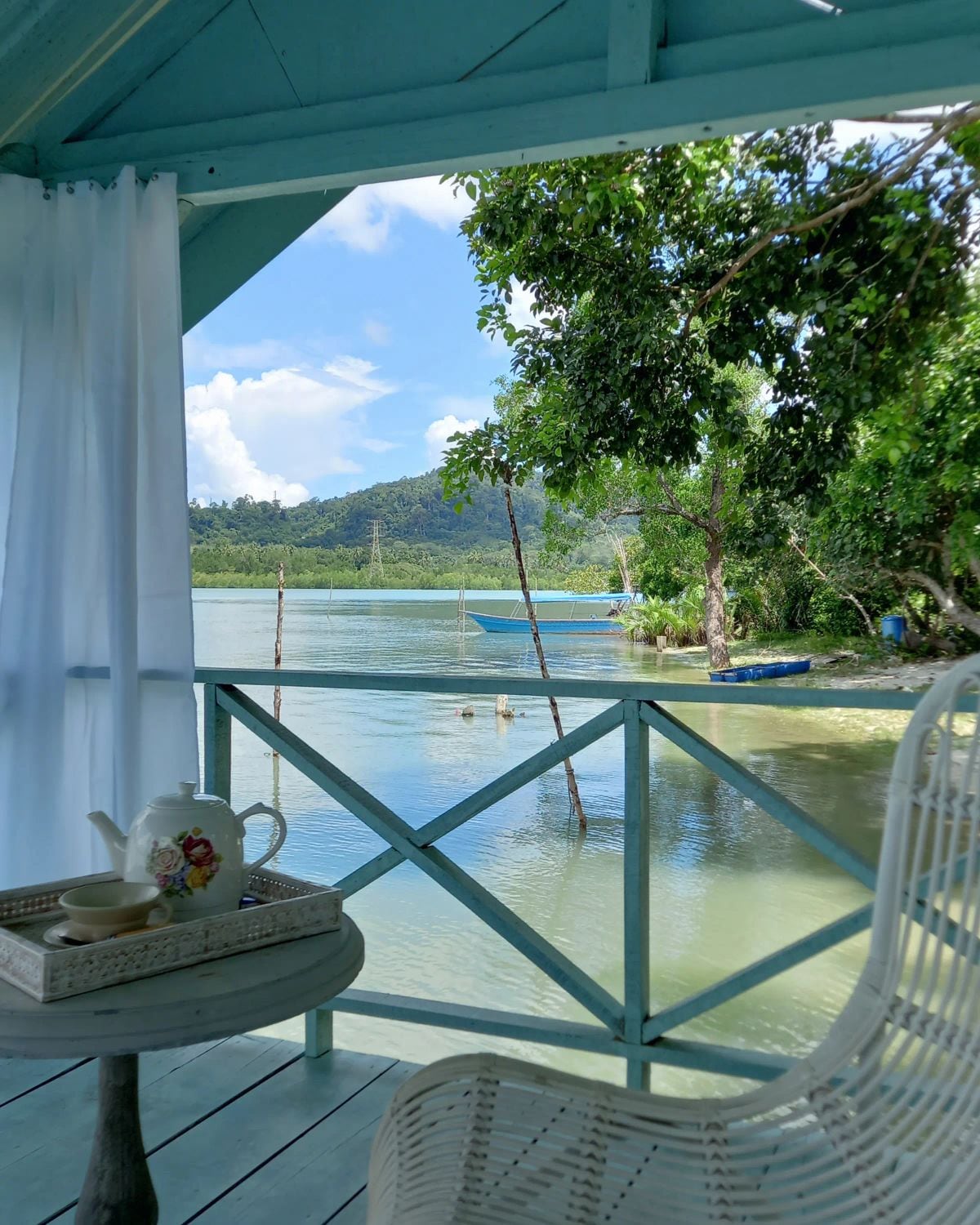 A serene view of a calm waterway is framed by a rustic porch, featuring a small table with a teapot and cup set. Lush greenery and tall trees provide shade, while a distant mountain line adds depth to the picturesque landscape.