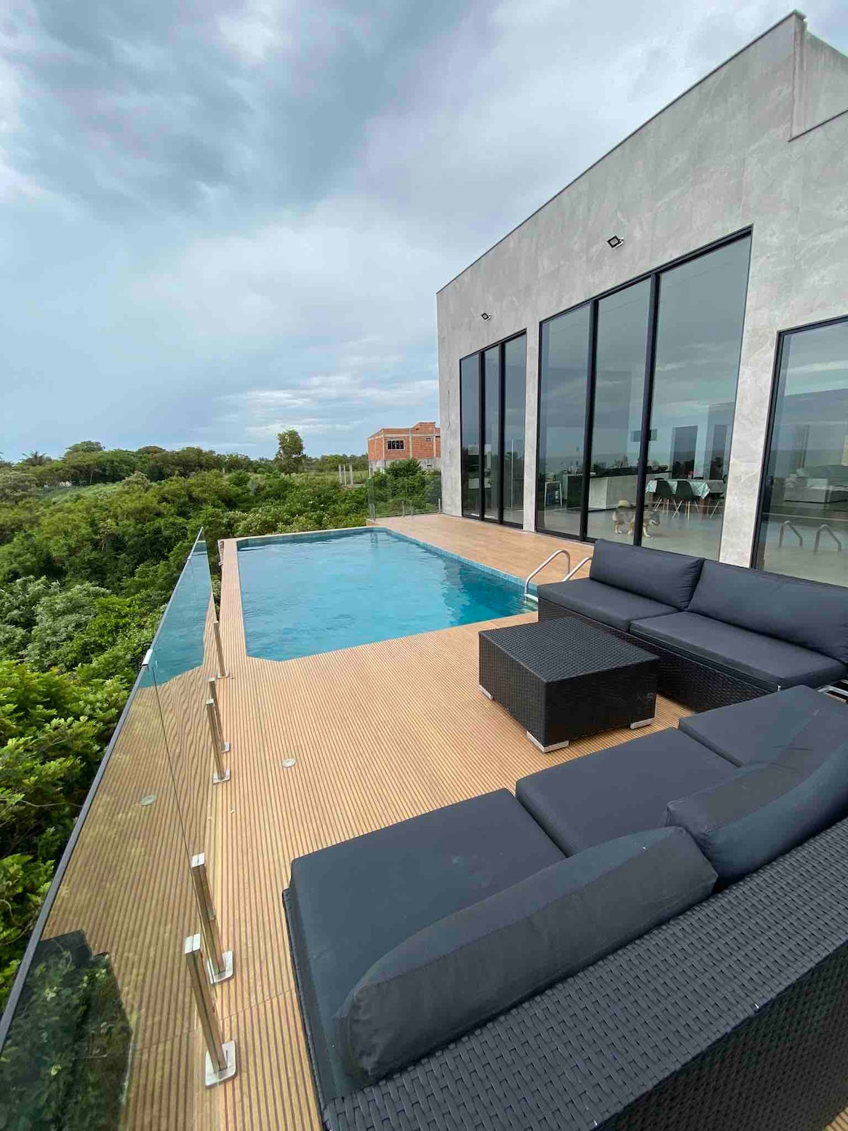 An infinity pool is seen extending towards the horizon, surrounded by a wooden deck. A seating area features dark outdoor sofas, offering a comfortable space to relax while enjoying the view of the greenery beyond.