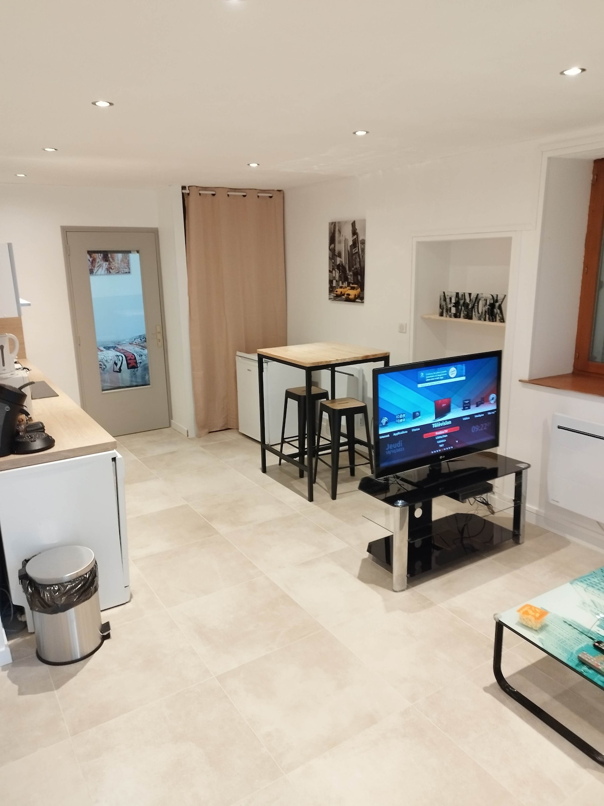 The living area is designed with modern amenities, featuring a sleek television on a glass stand. A small dining table with stools is positioned near the kitchen, complemented by minimalist cabinetry. Soft lighting highlights the tiled floor, creating a bright and spacious environment.