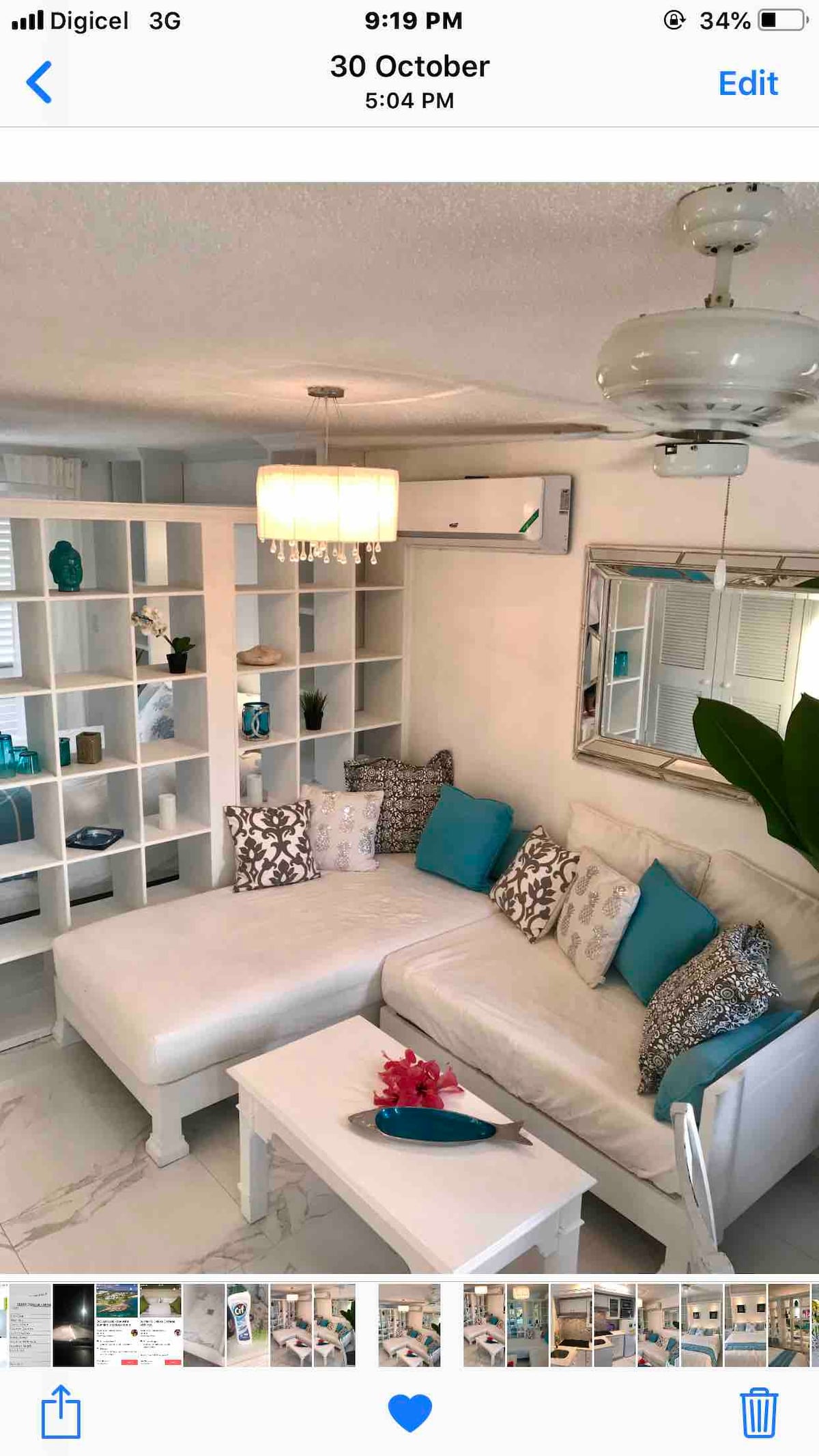 A bright and inviting living space features a white sectional sofa adorned with decorative pillows in shades of turquoise and neutral tones. A modern chandelier hangs overhead, while a shelf displays various decorative items. A large mirror reflects the light, enhancing the room's openness.