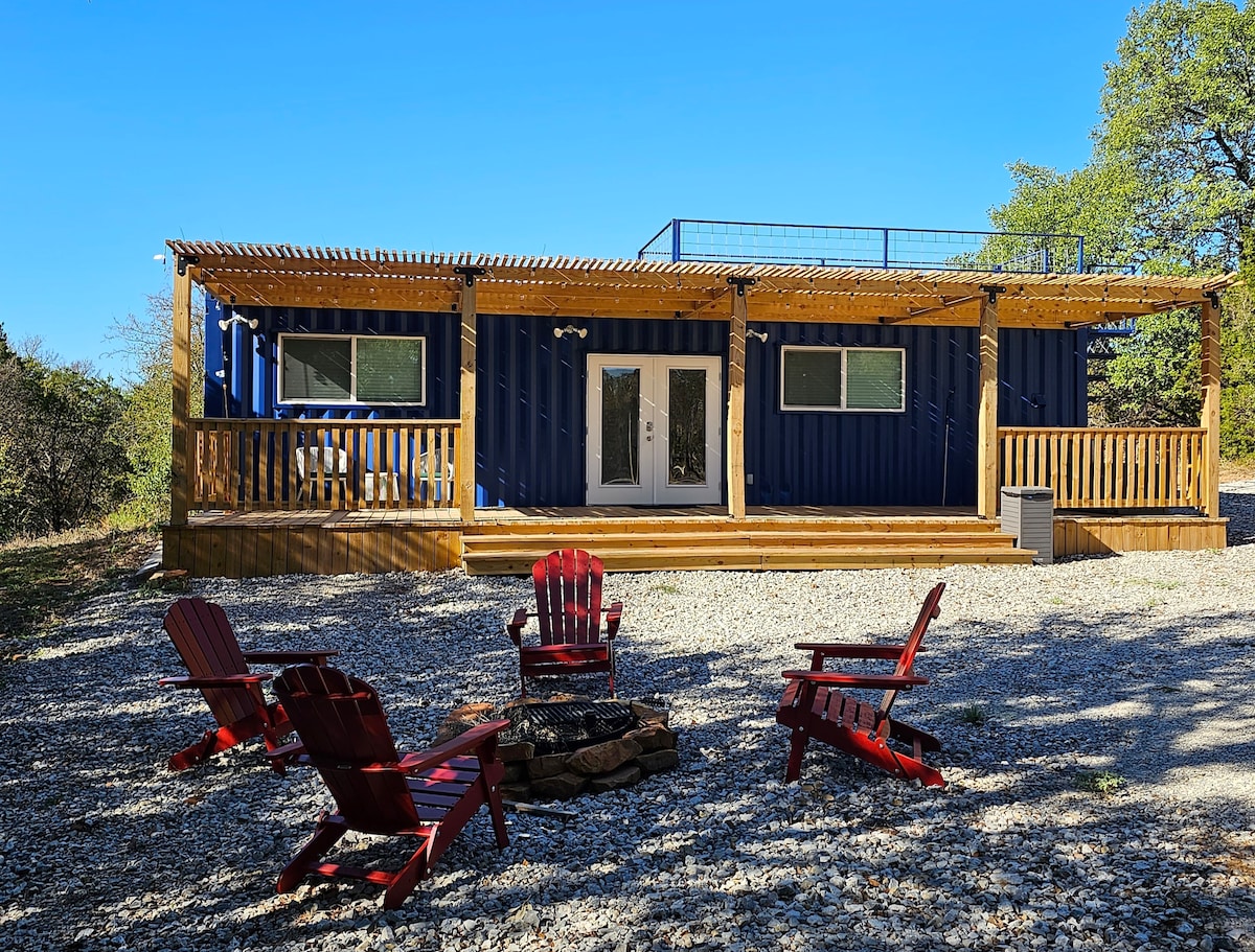 A vibrant blue container home is framed by a wooden deck and surrounded by gravel. A fire pit with red Adirondack chairs is positioned in front, inviting relaxation. The rooftop deck is visible above, providing additional outdoor space.