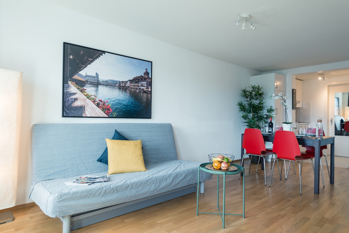A spacious living area features a gray sofa bed adorned with a yellow accent pillow. A round table set with four red chairs is positioned nearby. A vibrant artwork of the local scenery decorates the wall, while natural light brightens the wooden floor.
