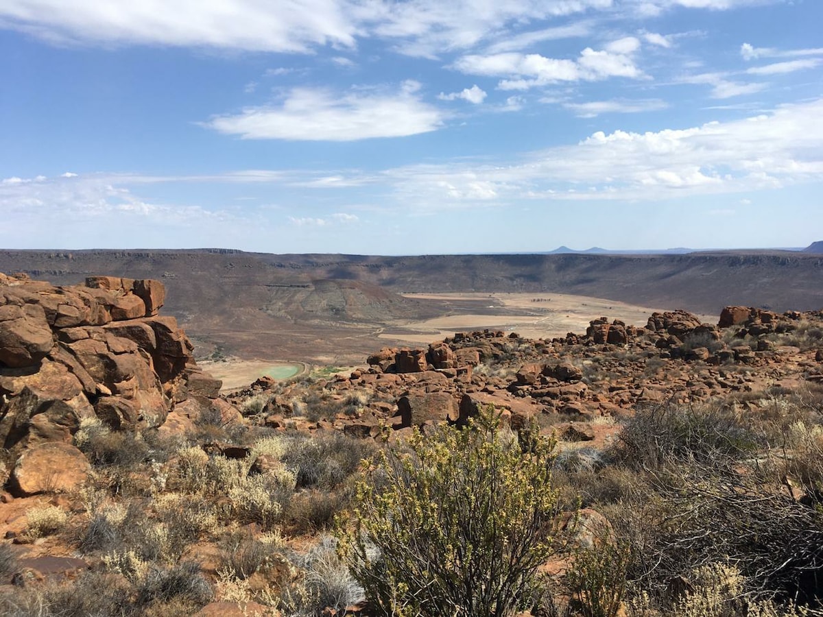 A panoramic view captures vast open plains framed by rugged mountains in the distance. Scattered shrubs and dry grasses dot the foreground, while rocky formations add texture to the landscape. The sky showcases a mix of clouds, hinting at a bright, sunny day.