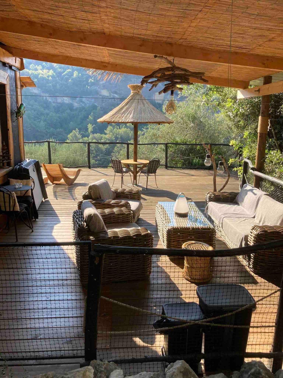 An inviting outdoor terrace is shown, featuring comfortable seating arrangements with wicker furniture. A round table is positioned under a thatched umbrella, while greenery surrounds the space. The wooden flooring and decorative elements contribute to a relaxed atmosphere in nature.