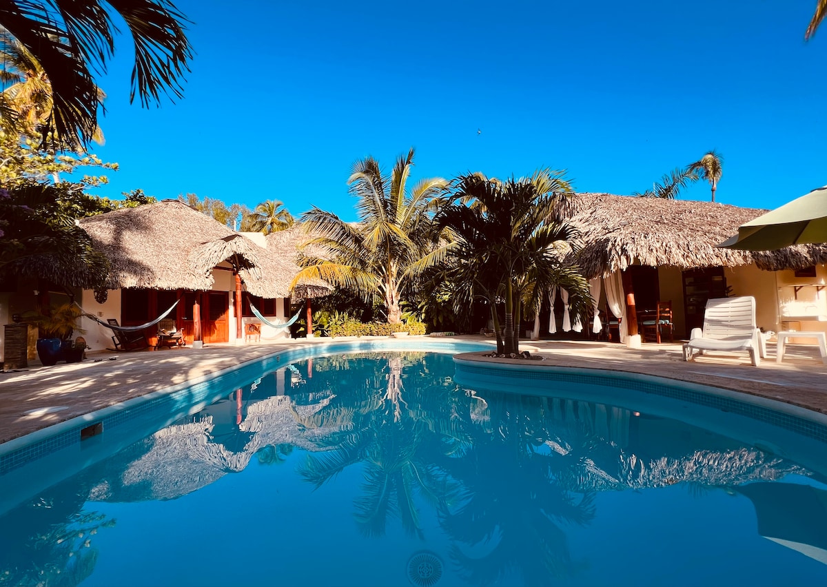 A tranquil saltwater pool reflects the clear blue sky, surrounded by lush gardens and palm trees. Thatched roof structures provide shaded lounging areas, enhancing the outdoor living experience. Comfortable seating and hammocks invite relaxation, creating a serene atmosphere for guests.