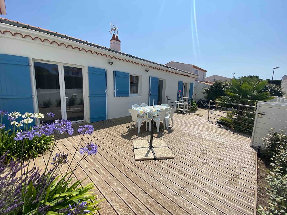 A sunlit wooden terrace is shown, featuring a dining table surrounded by white chairs. Bright blue shutters frame the house, and vibrant flowers are planted along the edges. The setting offers a spacious and inviting outdoor area for gatherings.