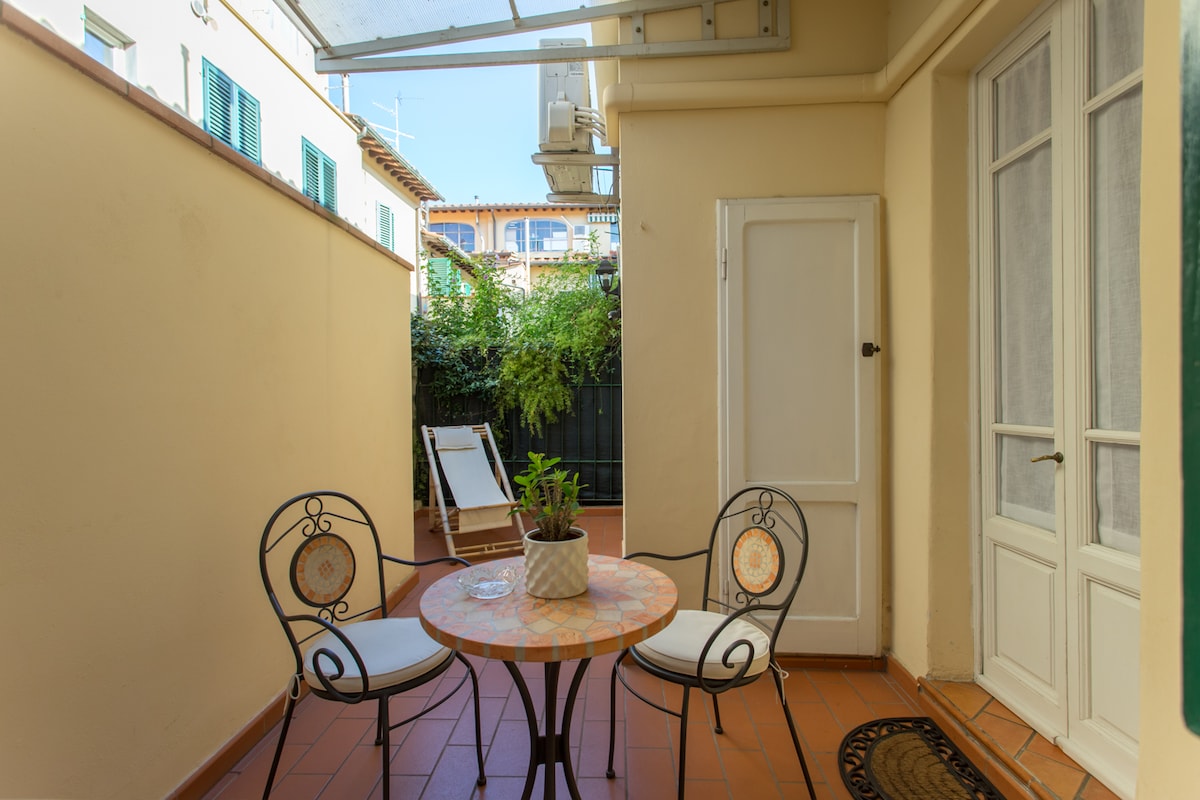 A private terrace is showcased with a round table and two chairs, featuring a decorative tile pattern. A potted plant sits center on the table. A sun lounger is visible in the background, surrounded by greenery, providing a relaxed outdoor space.