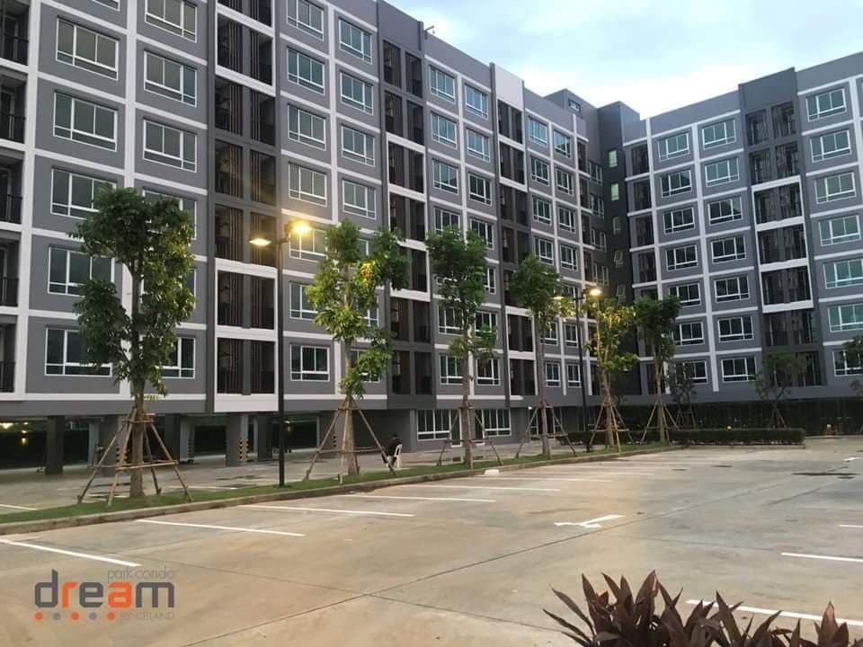 The building exterior features a modern design with multiple stories. Tree-lined walkways are visible, providing greenery and shade alongside a spacious parking area.