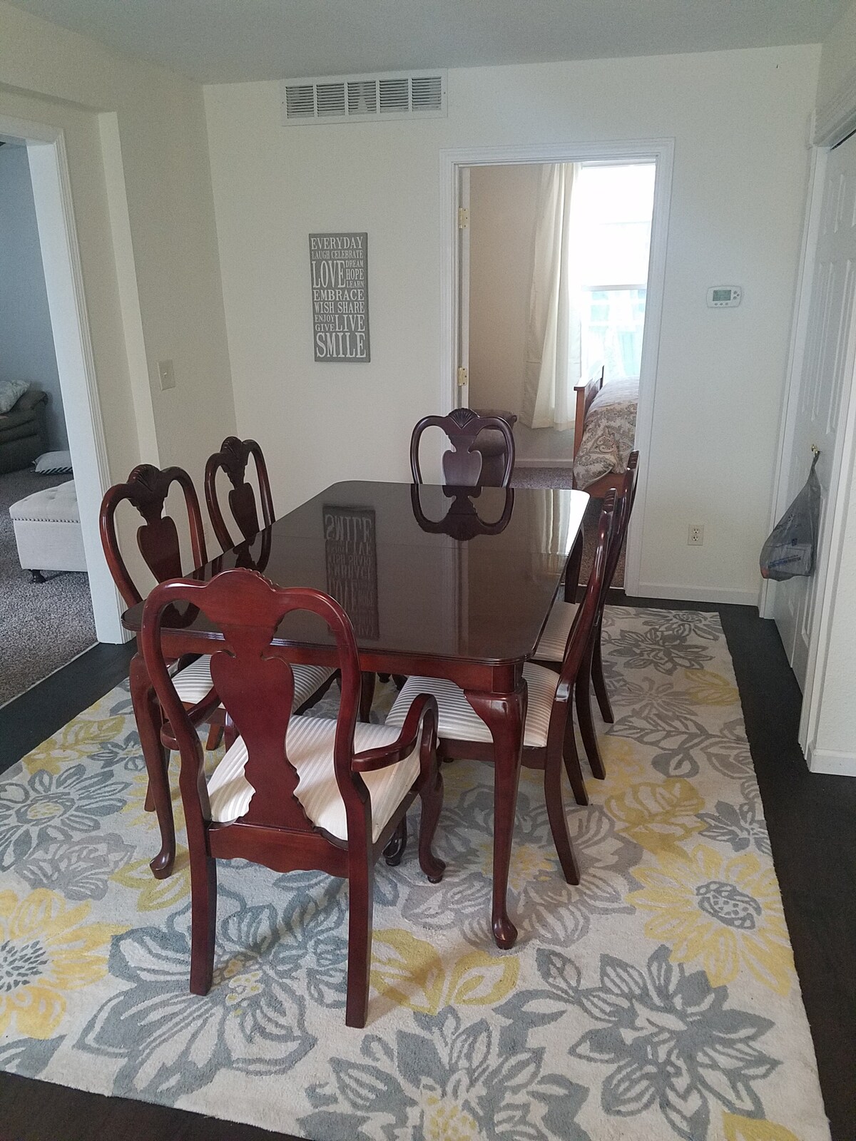 A traditional dining area features an elegant wooden dining table surrounded by six classic chairs. A patterned area rug in soft hues of grey and yellow lays beneath the table, complementing the light-colored walls. A doorway in the background leads to another room.