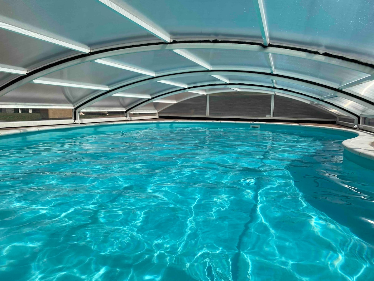 An indoor swimming pool is enclosed under a transparent arched roof, creating a bright and airy atmosphere. The water is clear and radiates a vibrant blue, with ripples reflecting soft light across the surface.