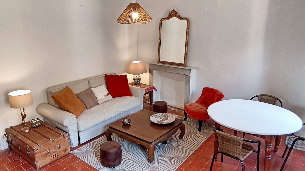 A spacious living area features a light-colored sofa adorned with colorful cushions. A wooden coffee table sits centrally, surrounded by seating options including upholstered chairs and stools. A decorative mirror hangs above a stone fireplace, with warm lamps providing additional lighting.