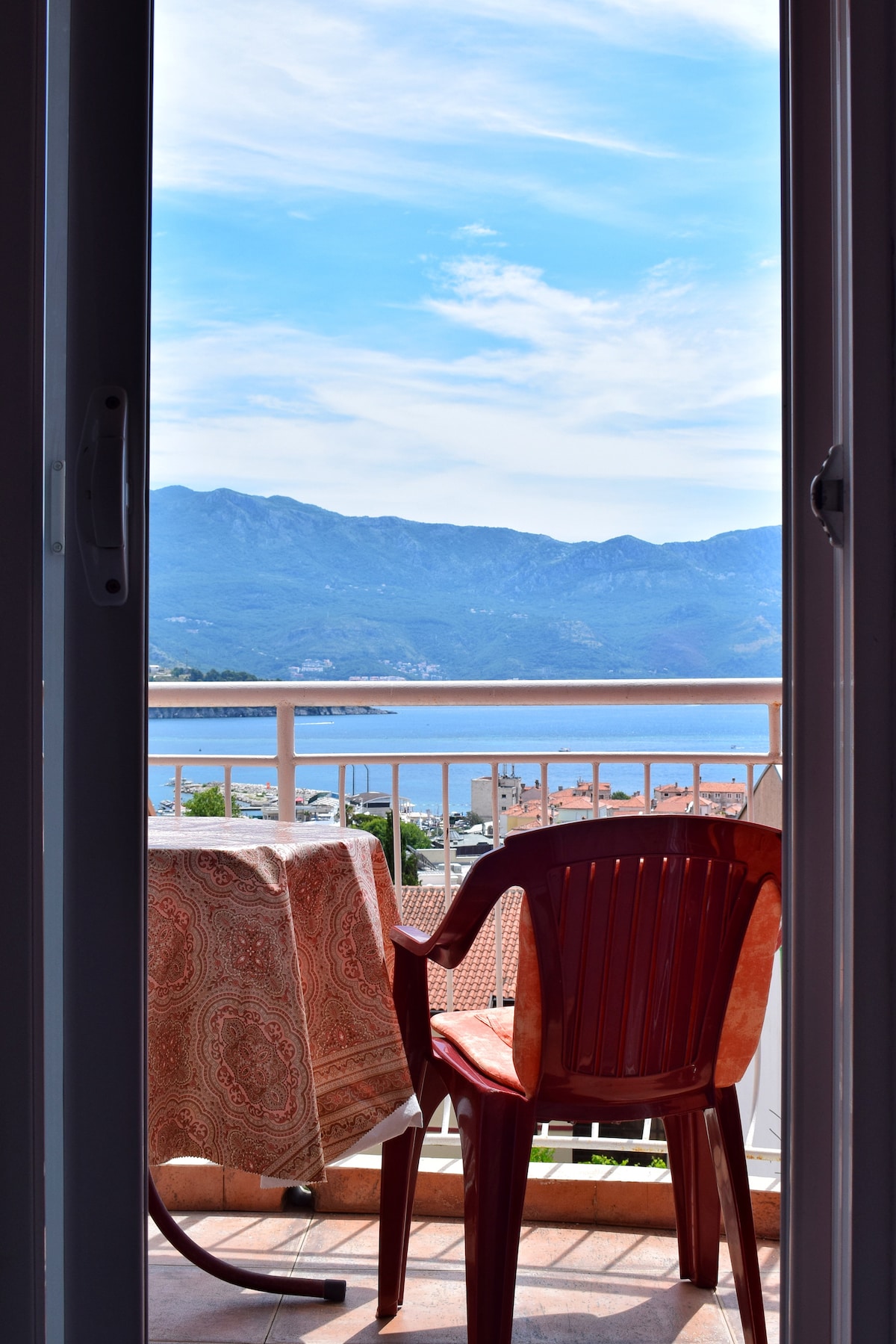 A balcony scene is shown with a red plastic chair and a small table covered by a patterned tablecloth. The view includes mountains and the sea, visible through the open sliding door, emphasizing a serene outdoor atmosphere.