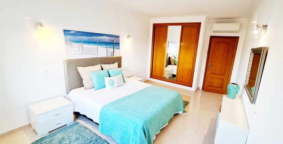 A bright bedroom features a spacious bed adorned with a light blue coverlet and pillows. A large mirror reflects the room, enhancing natural light. White nightstands and a plush rug complement the neutral decor, creating a serene atmosphere.