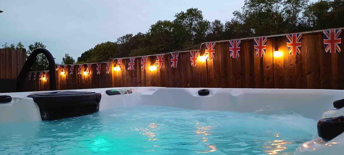 A relaxing outdoor jacuzzi is surrounded by wooden fencing adorned with string lights and decorative flags. The water is illuminated in a soft blue hue as dusk settles, creating a tranquil atmosphere ideal for unwinding.