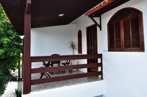 A private balcony is showcased with a wooden railing and a simple outdoor seating arrangement, including a small table and two chairs. The space is surrounded by greenery, with a large window allowing natural light into the adjoining area.